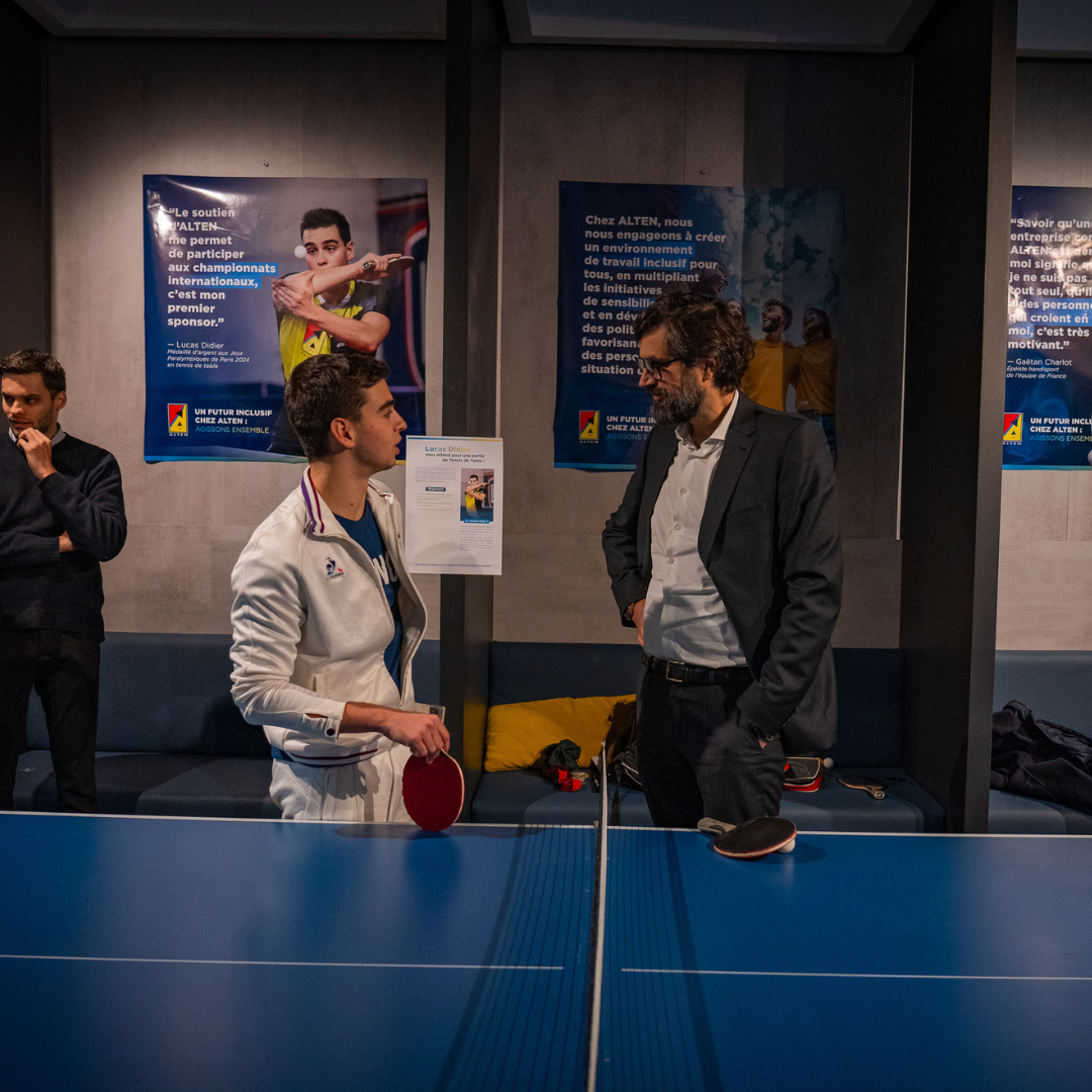 Lucas Didier (on the left), wearing sports attire and holding a table tennis paddle, talking with Pierre Bonhomme (on the right), dressed in a suit, in front of a ping-pong table during the handisport day organized by ALTEN at the Édouard Vaillant premises, with inclusive posters in the background.